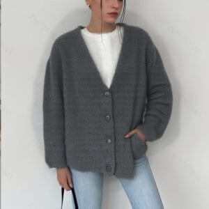 A woman wearing a gray cardigan and jeans, standing against a white wall with her arms crossed over her chest.