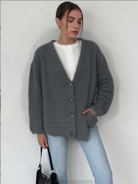 A woman wearing a gray cardigan and jeans, standing against a white wall with her arms crossed over her chest.