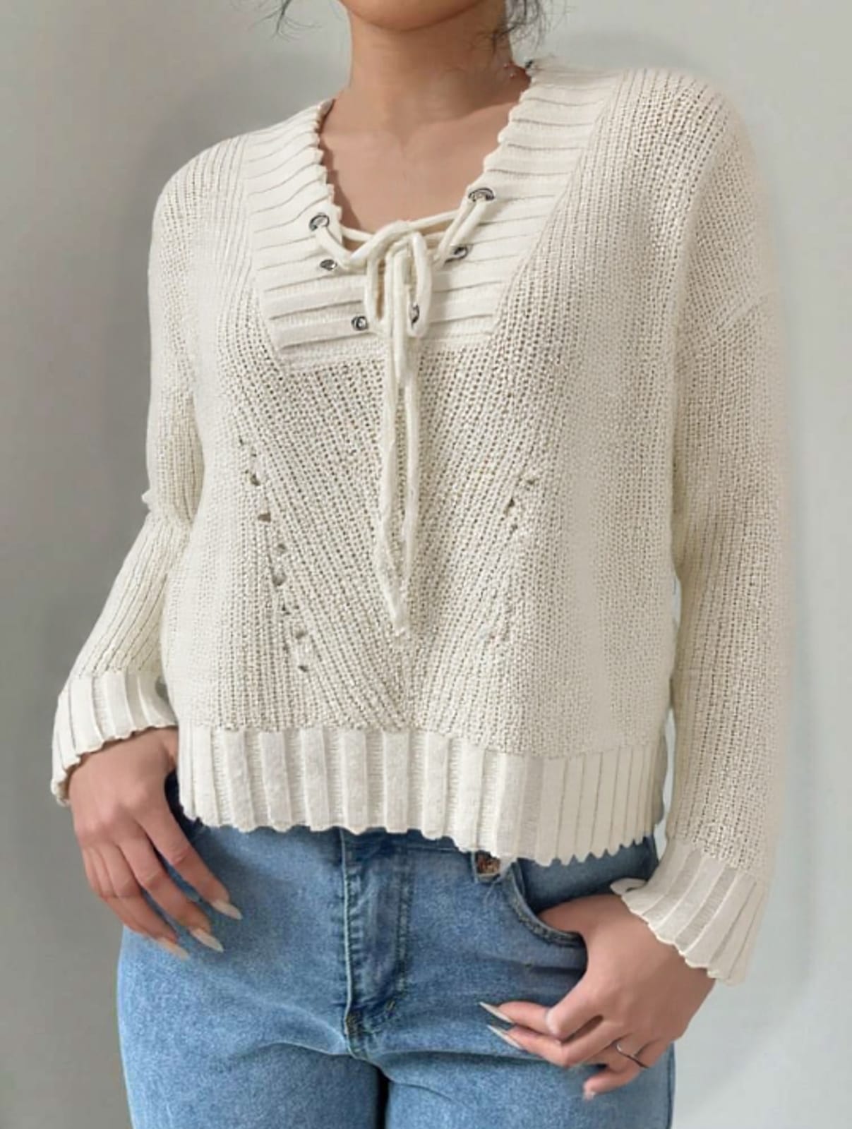 A person standing against a white background, wearing a white sweater with lace-up sleeves and blue jeans.