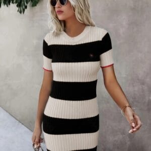 A woman with blonde hair is the main subject, standing against a gray wall while wearing a black and white striped dress.