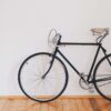 Minimalist black bicycle on hardwood floors against white wall with green plant leaves.