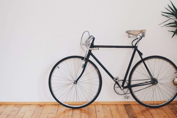 Minimalist black bicycle on hardwood floors against white wall with green plant leaves.