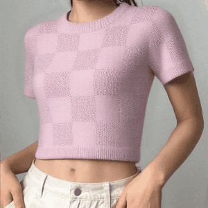 A person standing against a gray background, wearing a pink sweater with white checkerboard patterns and beige pants.