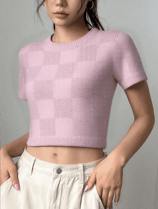 A person standing against a gray background, wearing a pink sweater with white checkerboard patterns and beige pants.