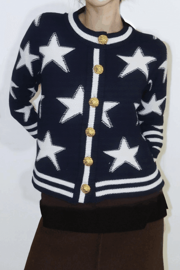 A person stands against a white background, wearing a navy blue sweater with white stars and gold buttons cinched at the.