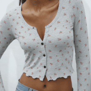 A woman stands against a white background, wearing a gray cardigan with orange flowers and blue jeans.