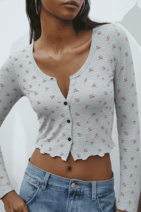 A woman stands against a white background, wearing a gray cardigan with orange flowers and blue jeans.