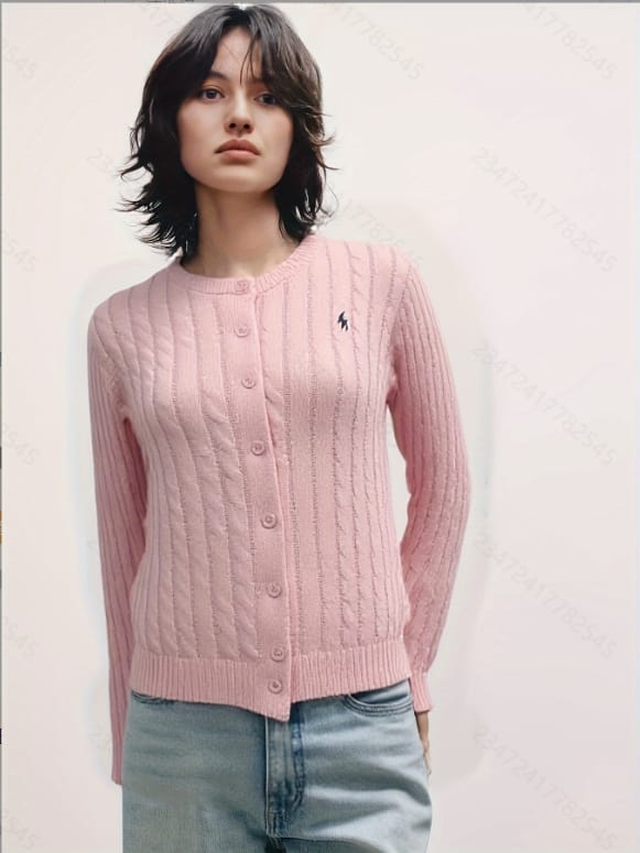 A young woman with short dark hair stands against a plain white background, wearing a pink cardigan over a white blouse and.