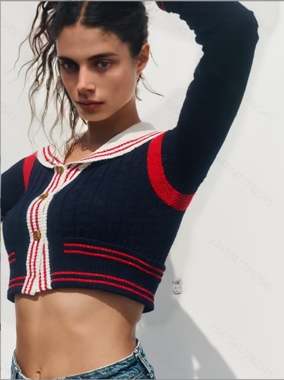 A young woman with long dark hair and bangs stands against a white background, wearing a navy blue sweater with red and.