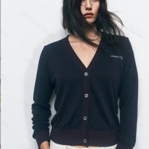 A woman standing against a white background, wearing a navy blue cardigan with a white logo and white pants.