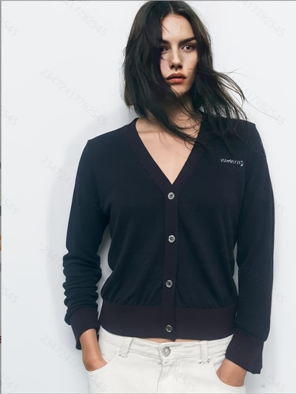 A woman standing against a white background, wearing a navy blue cardigan with a white logo and white pants.