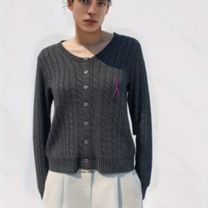 A woman stands against a white background, wearing a gray sweater with pink and purple accents, including a pink ribbon on.