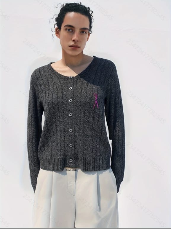 A woman stands against a white background, wearing a gray sweater with pink and purple accents, including a pink ribbon on.