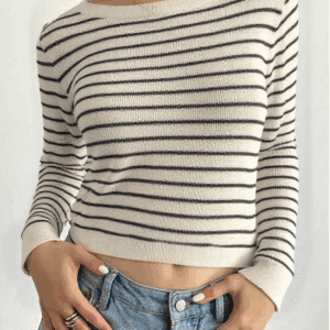 A person standing against a white background, wearing a beige and black striped sweater over blue jeans with gold rings.