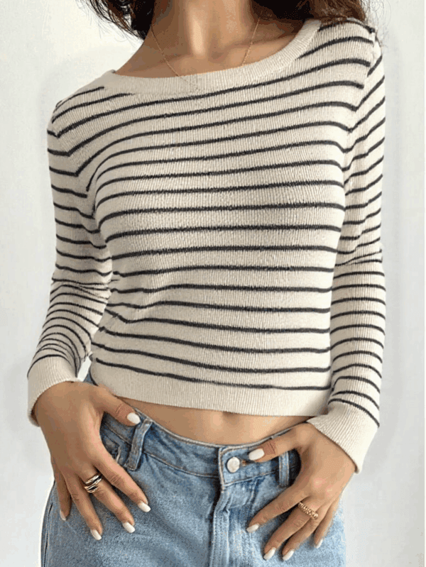 A person standing against a white background, wearing a beige and black striped sweater over blue jeans with gold rings.