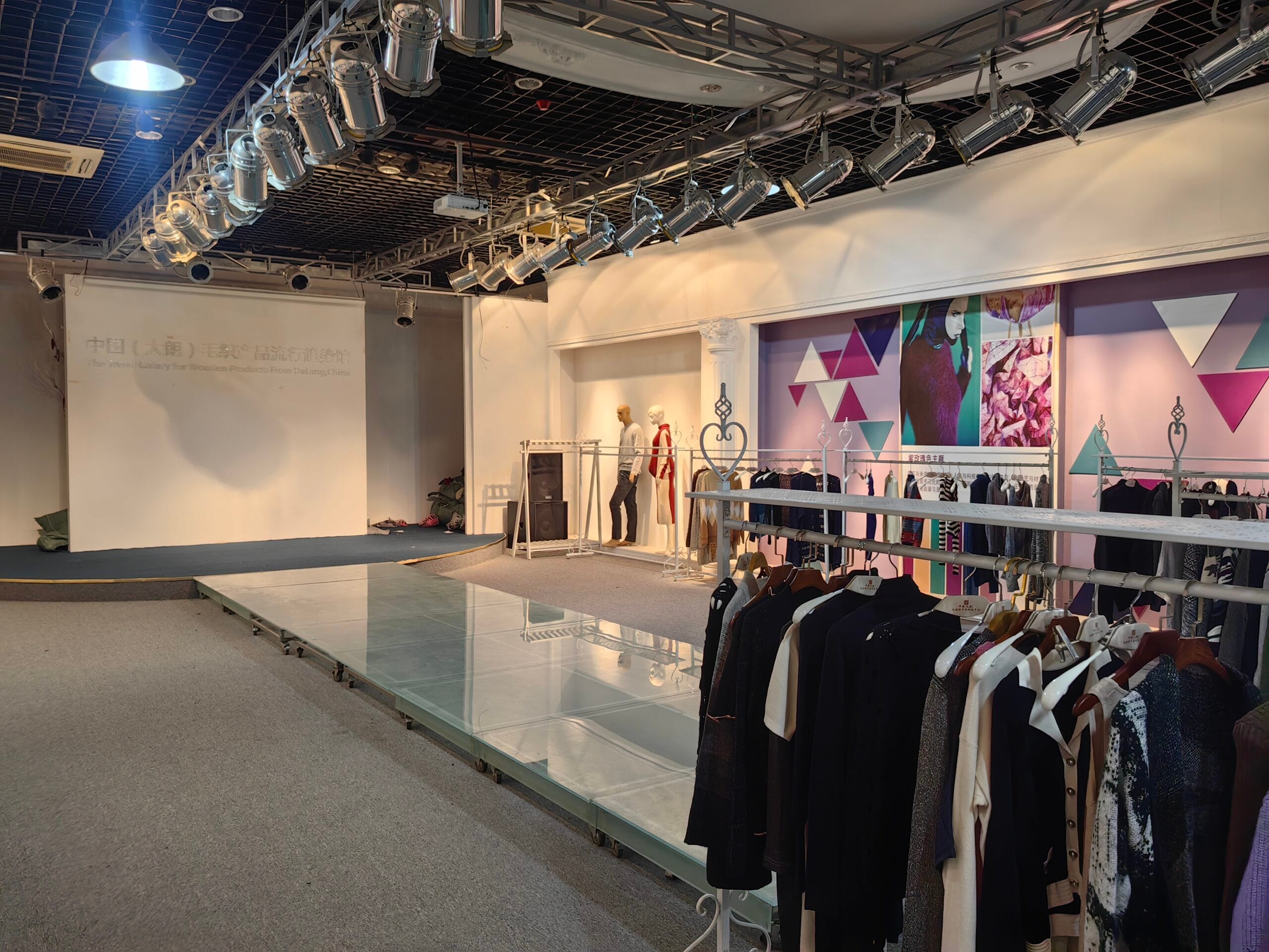 A large show room with a stage at one end of the room and racks of clothing hanging from a pole on the.