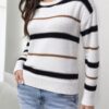A woman stands against a plain white wall, wearing a black and white striped sweater with a brown stripe down the center of.