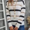 A woman stands against a gray wall, wearing a white and black striped sweater with a tassel necklace around her neck.