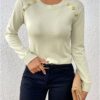 A woman wearing a white long-sleeved sweater with gold buttons and black pants, holding a beige clutch purse.