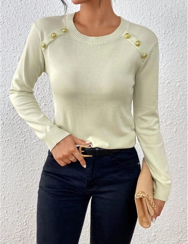 A woman wearing a white long-sleeved sweater with gold buttons and black pants, holding a beige clutch purse.