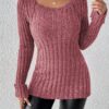 A woman stands confidently against a white wall, wearing a vibrant red sweater with a unique ribbed texture and a necklace.