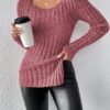 A woman stands against a white wall, wearing a pink sweater with a unique triangular neckline and black pants that match the.