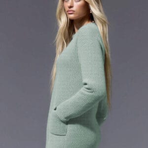 A woman with long blonde hair stands against a gray background, wearing a light green sweater and blue jeans.