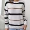 A woman stands against a white wall, wearing a gray and black striped sweater with black horizontal stripes.