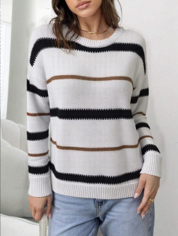 A woman stands against a white wall, wearing a gray and black striped sweater with black horizontal stripes.