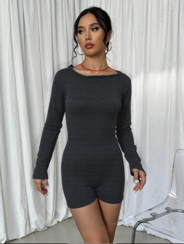 A woman stands confidently against a white curtain backdrop, wearing a black jumpsuit and red necklace that catches the eye.