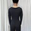 In the center of a room with white walls and a gray curtain, a person stands wearing a black jumpsuit or one-piece swimsuit.