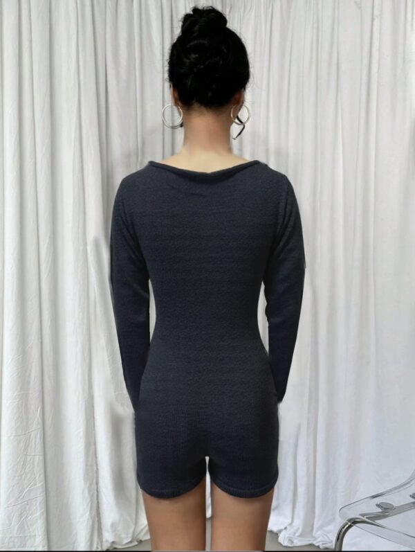 In the center of a room with white walls and a gray curtain, a person stands wearing a black jumpsuit or one-piece swimsuit.