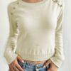 A woman wearing a beige sweater with gold buttons and blue jeans against a white background.