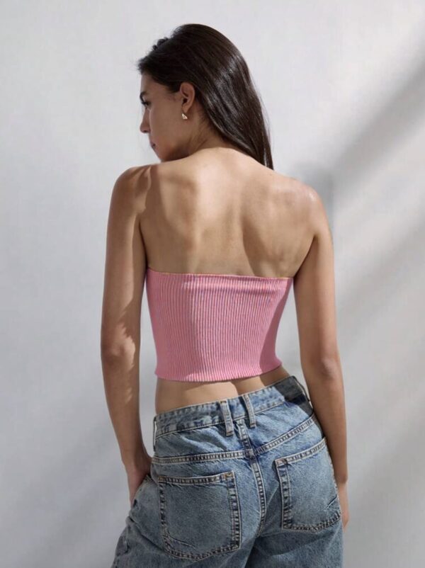 A woman standing against a plain white wall, wearing a pink crop top and blue jeans.