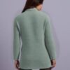 A woman is standing against a purple background, wearing a light green sweater with a ribbed texture and black pants.