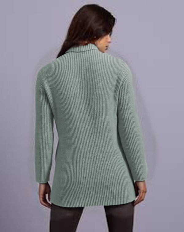 A woman is standing against a purple background, wearing a light green sweater with a ribbed texture and black pants.