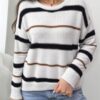 A woman stands against a white wall, wearing a white and black striped sweater with brown stripes.