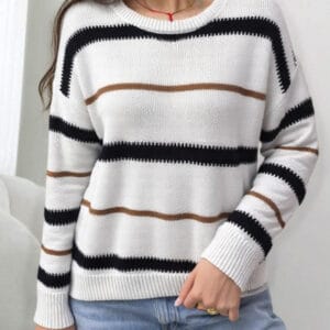 A woman stands against a white wall, wearing a white and black striped sweater with brown stripes.