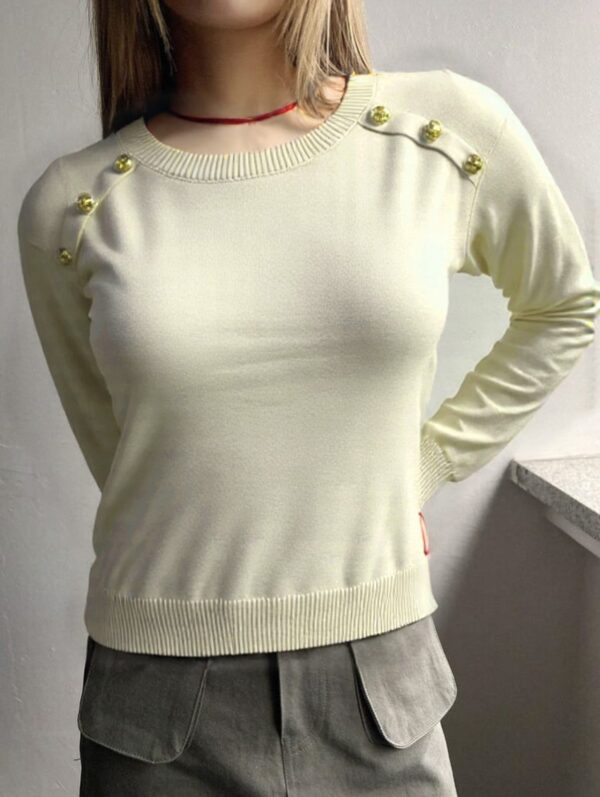 A person standing against a white background, wearing a beige sweater with gold buttons and gray pants.