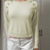 A person standing against a white wall, dressed in a beige sweater and white pants with red accents.